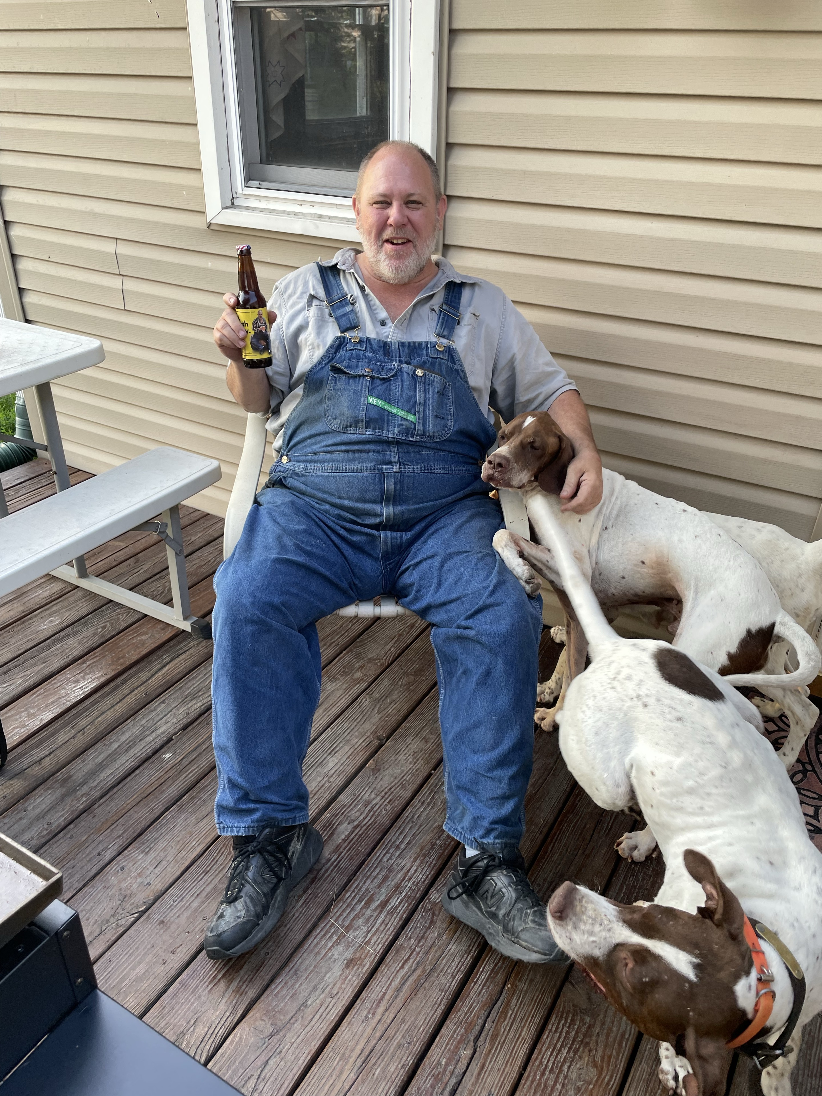 Mike Pinkerton in his overalls, holding his home-brew on the porch with his dogs.